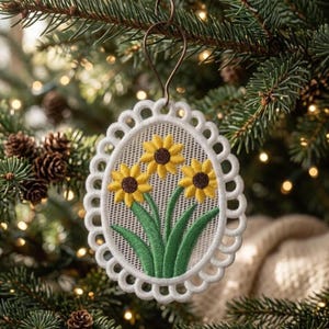 Może przedstawiać: Owalna ozdoba choinkowa z haftowanym wzorem trzech słoneczników z żółtymi płatkami i brązowymi środkami, na tle zielonych liści. Ozdoba ma białą, falistą obwódkę i jest zawieszona na metalowym haczyku.