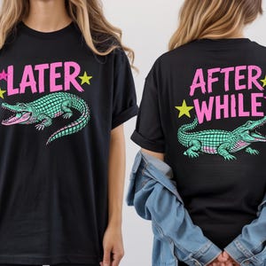 May include: Black t-shirt featuring a teal and pink alligator graphic with the text "LATER" on the front and "AFTER WHILE" on the back. The design includes pink stars and yellow stars. The shirt is a casual, unisex style.