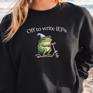 Könnte beinhalten: Schwarzes Sweatshirt mit dem Text "Off to write IEPs". Die Grafik zeigt einen grünen Frosch mit Zaubererhut, der Roller fährt. Der Frosch hat große Augen und lächelt. Das Sweatshirt hat eine einfarbige schwarze Farbe.