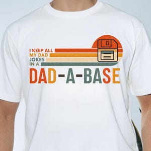 Könnte beinhalten: Weißes T-Shirt mit Retro-Design, das eine Diskettengrafik und den Text "I KEEP ALL MY DAD JOKES IN A DAD-A-BASE" zeigt. Das Design umfasst orange, gelbe und grüne Streifen.