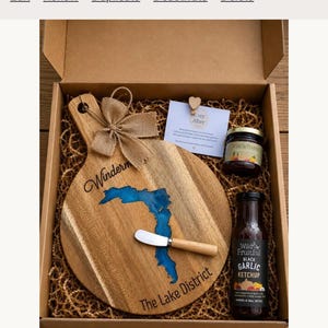 Puede incluir: Una caja de regalo que contiene una tabla de servir redonda de madera con una incrustación de resina azul con la forma de Windermere, un pequeño esparcidor de madera, un tarro de conserva y una botella de kétchup de ajo negro Wild & Fruitful.