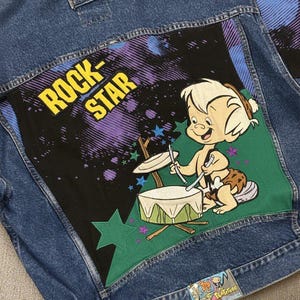 May include: A blue denim jacket with a cartoon graphic on the back. The graphic shows a character playing drums, with the words "ROCK-STAR" in yellow and black, and purple and green details.