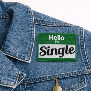 May include: A green and white embroidered patch with the text "Hello I'm Single" in black and white lettering, attached to a blue denim jacket. The patch has a name tag design.