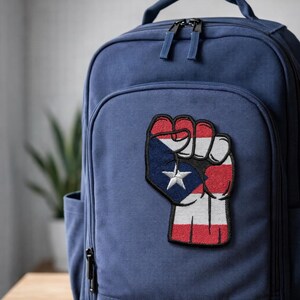 May include: A navy blue backpack with a patch featuring a clenched fist design. The patch incorporates the colors and star from the Puerto Rican flag. The backpack has a front pocket, two zippers, and a top handle.