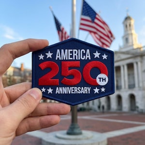 May include: A hexagonal embroidered patch with a dark blue background. The text reads AMERICA 250TH ANNIVERSARY in white, with the number 250 in large red font. Four white stars are positioned above and below the text on the left and right sides.