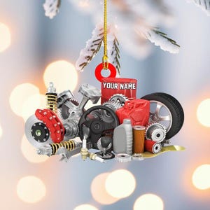 May include: A Christmas ornament featuring an assortment of car parts, including a tire, steering wheel, and engine components. A red can with the text "YOUR NAME" is also included. The ornament is suspended by a gold string.