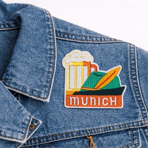 May include: Embroidered patch featuring a beer stein, a traditional hat with a feather, and the word "MUNICH" in orange. The patch is attached to a blue denim jacket, showcasing a casual style.