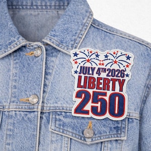May include: A light blue denim jacket with a patriotic patch. The patch features the text "JULY 4TH 2026 LIBERTY 250" in red and blue, with fireworks and stars. The jacket has silver buttons.