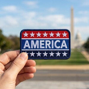 May include: A rectangular embroidered patch featuring a red top section with seven white stars, a blue middle section with the word AMERICA in white capital letters, and a dark blue bottom section with seven white stars, all with a dark blue border.