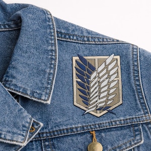May include: A denim jacket with a patch featuring a stylized emblem of blue and white wings on a beige background. The jacket has a classic collar and button closure.