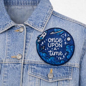 May include: A light blue denim jacket with a round, blue embroidered patch. The patch features a rocket ship, a flying saucer, stars, and the words "once upon a time" in white script. The jacket has silver buttons.