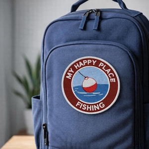 May include: A blue backpack with a circular patch that reads "My Happy Place Fishing." The patch features a red and white fishing bobber over blue water. The backpack has a top handle and two zippered compartments.