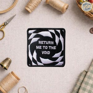 May include: Square black and white embroidered patch with the text "RETURN ME TO THE VOID". The patch features a geometric design in white against a black background. The patch is surrounded by sewing supplies.