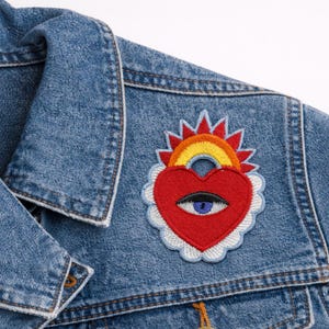 May include: A denim jacket with a colorful embroidered patch. The patch features a red heart with an eye, a rainbow, and a sun design. The patch is surrounded by a white and light blue scalloped border. The jacket is a light blue wash.
