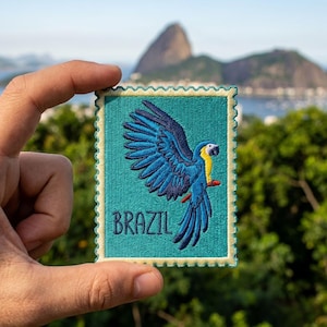 May include: A rectangular embroidered patch with a scalloped edge, featuring a teal background, a blue and yellow macaw parrot in flight, and the word BRAZIL stitched in dark blue thread at the bottom left corner of the design.
