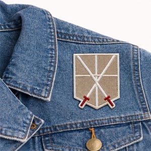 May include: Embroidered patch featuring a beige shield design with two crossed white swords and red accents. The patch is sewn onto a blue denim jacket, showcasing a classic and durable aesthetic.