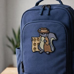 May include: A navy blue backpack with a cartoon patch of a squirrel wearing a fedora and trench coat, holding a stack of what appears to be money. The patch is embroidered with brown, gray, and tan colors. The backpack has a top handle and two zipper pulls.