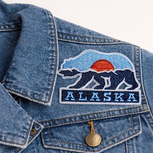 May include: A blue denim jacket with a decorative patch featuring a bear silhouette, a red sun, and the word "ALASKA" in white letters. The patch is embroidered with blue and white thread.