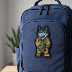 May include: A navy blue backpack with a patch of a wolf wearing a military uniform. The patch is embroidered with shades of gray, tan, and green. The wolf has blue eyes and is holding a rifle.