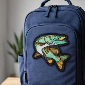 May include: A navy blue backpack with a large embroidered patch of a fish. The patch features a green and yellow fish with a black border. The backpack has a front pocket and two zippers.