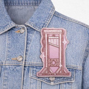 May include: A light blue denim jacket with a pink guillotine patch. The patch is embroidered with a detailed design of the guillotine, featuring a pink background and a darker pink outline. The jacket has silver buttons.