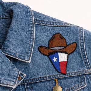 May include: A denim jacket with a brown cowboy hat patch featuring the Texas state flag. The patch is a combination of blue, white, and red, with a white star. The jacket has a classic collar and buttoned cuffs.