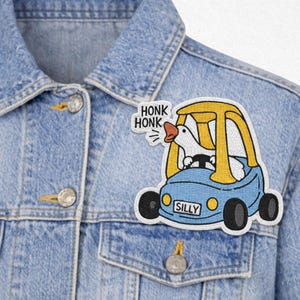 May include: Embroidered patch featuring a white goose driving a blue car with yellow accents. The patch has the word "SILLY" on the car and "HONK HONK" on a separate tag. The patch is attached to a denim jacket.