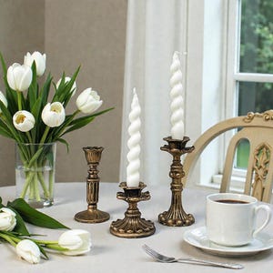 Könnte beinhalten: Ein Stillleben mit weißen Spiralkerzen in verzierten goldenen Kerzenhaltern, einem Strauß weißer Tulpen in einer Glasvase und einer Tasse Kaffee auf einer weißen Untertasse. Eine Gabel liegt auf dem Tisch.