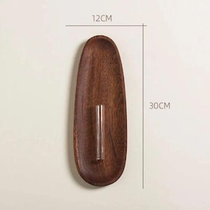 Pu&ograve; includere: Un vassoio ovale in legno marrone scuro, largo 12 cm e lungo 30 cm, con un piccolo tubicino di vetro trasparente al centro. Il vassoio ha una finitura liscia e lucida, su sfondo neutro.
