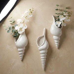 May include: Three white ceramic seashell-shaped wall vases, each with a different design. Two vases hold white flowers and green foliage. The vases are mounted on a beige wall, creating a coastal theme.