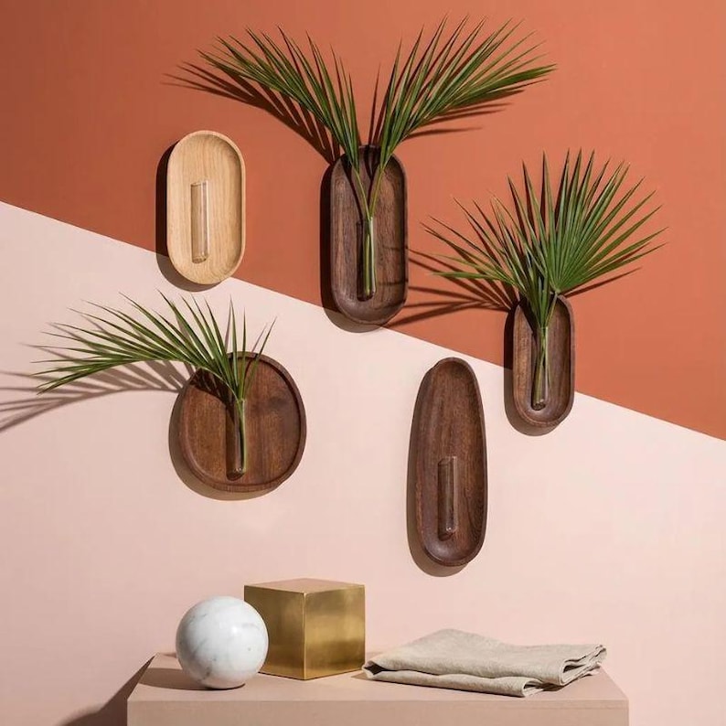 Pu&ograve; includere: Una collezione di vasi da parete in legno di varie forme ovali e circolari, ciascuno con un tubicino di vetro con fronde di palma verdi. I vasi sono montati su una parete bicolore rosa e arancione. Una sfera di marmo, un cubo d'oro e un panno di lino sono su una mensola.