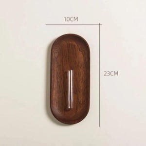 Pu&ograve; includere: Un vaso da parete in legno, lungo 23 cm e largo 10 cm, con un tubo di vetro trasparente per esporre fiori. Il vaso ha una finitura marrone scuro e una forma ovale, perfetto per l'arredamento minimalista.