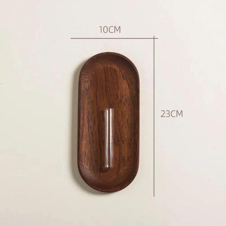 Pu&ograve; includere: Un vaso da parete in legno, lungo 23 cm e largo 10 cm, con un tubo di vetro trasparente per esporre fiori. Il vaso ha una finitura marrone scuro e una forma ovale, perfetto per l'arredamento minimalista.