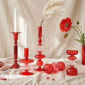 Può includere: Una collezione di portacandele e vasi in vetro rosso. L'immagine presenta varie forme e dimensioni, tra cui candelabri alti e sottili e elementi decorativi sferici. Candele bianche e fiori rossi creano contrasto.