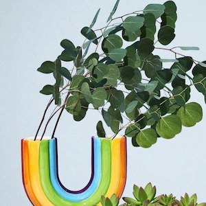 May include: A colorful, rainbow-shaped ceramic vase holds green eucalyptus branches. The vase features bands of yellow, orange, green, blue, and purple. Small succulents are arranged in front of the vase, creating a vibrant display.