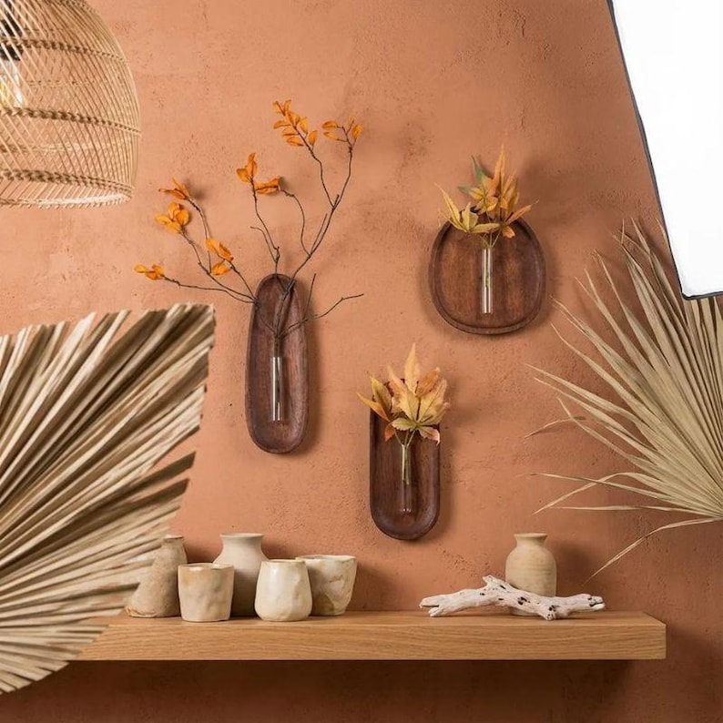 Pu&ograve; includere: Tre vasi da parete in legno con tubi di vetro, ciascuno contenente fiori secchi e rami. I vasi sono montati su una parete color terracotta. Una mensola in legno sottostante espone vasi in ceramica e un pezzo di legno trasportato dalla corrente.