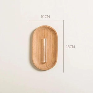 Pu&ograve; includere: Un portavaso ovale in legno con un tubo di vetro trasparente per un singolo stelo. Il supporto &egrave; largo 10 cm e alto 18 cm. La venatura naturale del legno &egrave; visibile. Design minimalista.