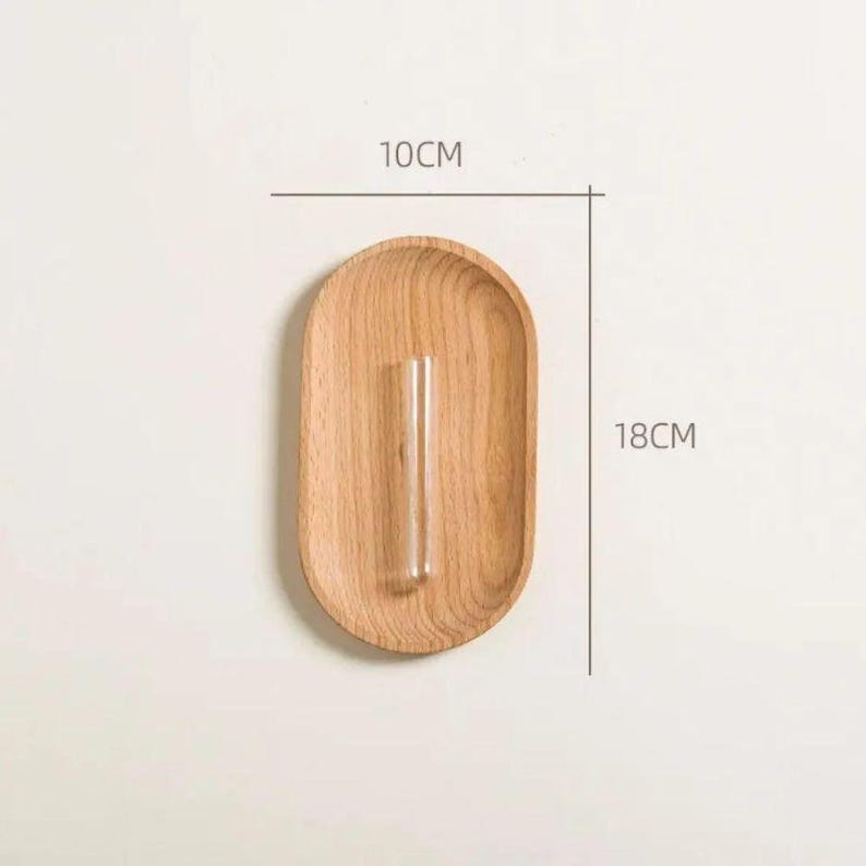 Pu&ograve; includere: Un portavaso ovale in legno con un tubo di vetro trasparente per un singolo stelo. Il supporto &egrave; largo 10 cm e alto 18 cm. La venatura naturale del legno &egrave; visibile. Design minimalista.