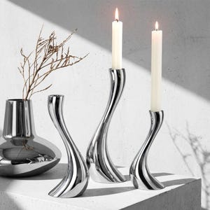 Può includere: Una collezione di elementi decorativi per la casa moderni, color argento. L'allestimento comprende tre portacandele ondulati con candele bianche accese e un vaso con rami secchi. Gli oggetti sono esposti su una superficie bianca.