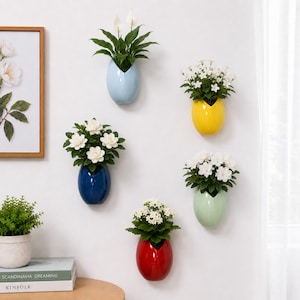 May include: A selection of wall-mounted ceramic planters, each filled with green plants and white flowers. The planters are in various colours, including blue, yellow, green, and red. The display is set against a white wall. A framed floral print and books are visible.