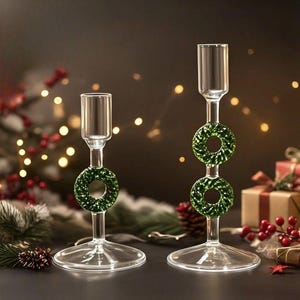 Puede incluir: Dos candelabros de vidrio transparente con diseños de coronas verdes. Los candelabros tienen una base ancha y un tallo delgado. Las coronas están hechas de pequeños materiales verdes texturizados. El fondo es oscuro con luces borrosas y decoraciones.