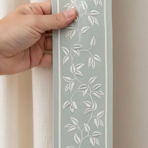 May include: A decorative fabric trim featuring a light green background with a white floral vine pattern. The trim is bordered by thin white lines. The trim is held up against a cream-colored curtain.