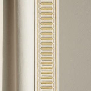 May include: A close-up of a cream-coloured curtain with a decorative border. The border has a repeating geometric pattern in gold and cream, adding elegance to the fabric. The curtain's texture is visible.