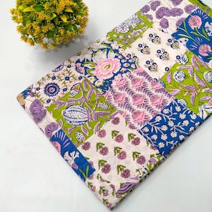 Pode incluir: Um tecido dobrado com um design de patchwork com padrões florais em rosa, azul, verde e roxo. O tecido tem um fundo branco e está ao lado de uma pequena planta redonda amarela.