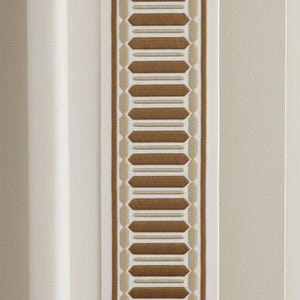 Könnte beinhalten: Vertikales Bild eines dekorativen Zierbands mit einem sich wiederholenden geometrischen Muster in Braun- und Beigetönen. Das Zierband wird von weißem Stoff begrenzt und vor einem neutralen Hintergrund platziert.