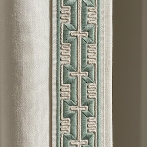 Può includere: Primo piano di un tessuto color crema con un bordo decorativo. Il bordo presenta un motivo ripetuto di croci e forme geometriche in verde e bianco sporco. Il tessuto ha un aspetto strutturato.