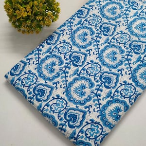 May include: Folded fabric with a white background and a repeating pattern of blue floral designs. The floral pattern includes paisley and heart-shaped elements in varying shades of blue. A small, yellow-green plant is in the upper left corner.