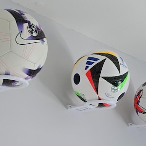 Op de afbeelding: Drie voetballen gemonteerd op een witte muur. De ballen hebben verschillende ontwerpen, waaronder een Nike-logo en kleurrijke patronen. Elke bal wordt vastgehouden door een witte, cirkelvormige houder die aan de muur is bevestigd.