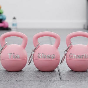 May include: Three light pink kettlebell-shaped keychains with silver metal rings. Each keychain features a custom name engraved in silver: Tia, Shep, and Charlie. One keychain includes a small beige tag with a kettlebell icon and the text PLA Ring Safe.