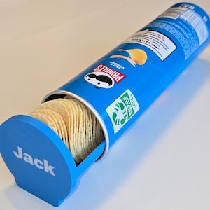 May include: A blue Pringles tube with the name "Jack" on the lid, partially open to show potato crisps. The Pringles logo and "Salt & Vinegar" are visible. The container is recyclable. The crisps are golden brown.